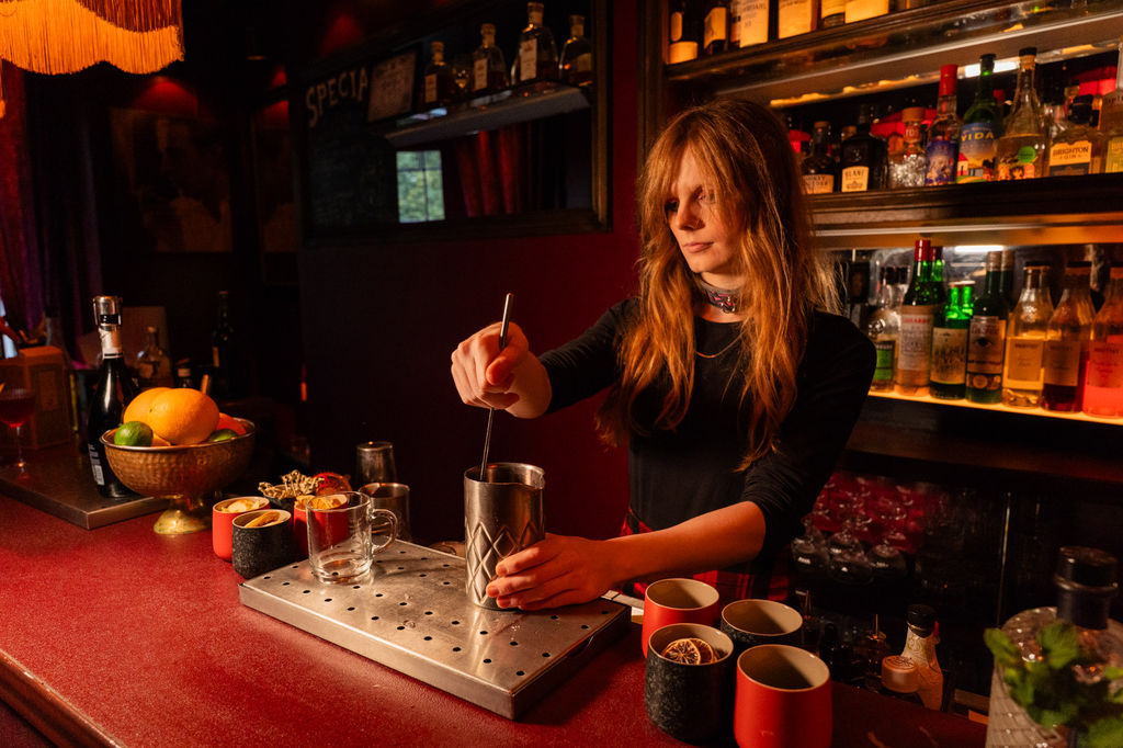 A lady making a cocktail in a red painted bar with a bowl of fresh citrus fruit at the end of the bar.