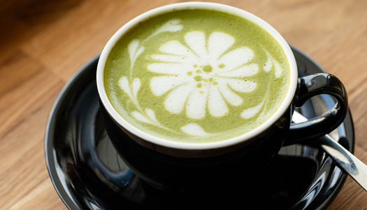 Green frothy matcha latte with flower latte art in a black mug. Headlining image to our Matcha Brighton guide