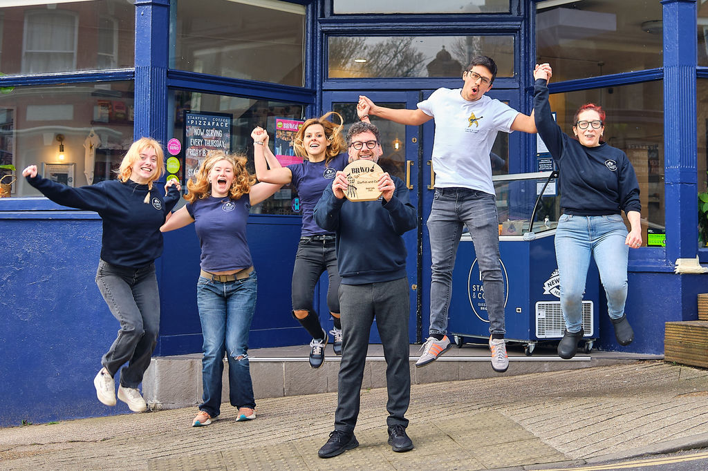 The team celebrating their BRAVO win in front of the blue exteriors of the cafe.