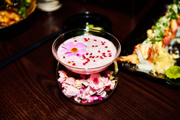 a cocktail is centre of the picture. It's a pretty bowl of blossoms with a pink foamy cocktail topped with a flower and red sprinkles. Plates of sushi are out of focus in the back of the picture