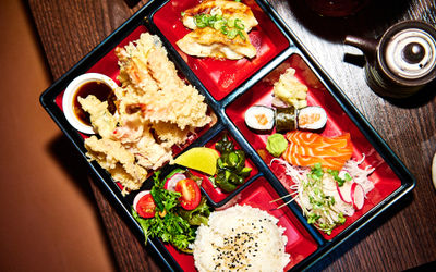 Colourful Japanese bento box with tempura prawns and dipping sauce, pan-fried gyoza, salmon sashimi and maki, rice with sesame, salad and pickles