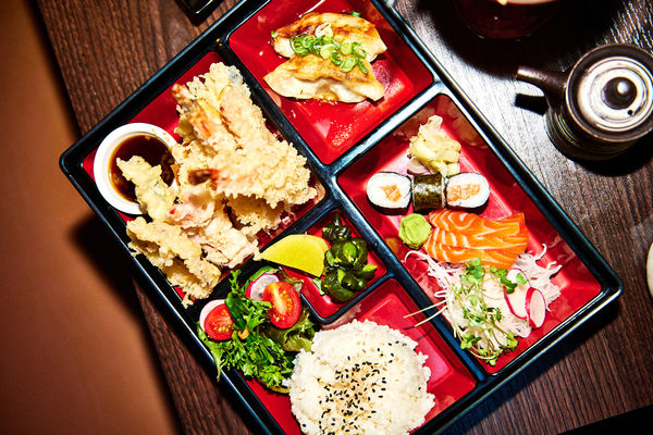 Colourful Japanese bento box with tempura prawns and dipping sauce, pan-fried gyoza, salmon sashimi and maki, rice with sesame, salad and pickles