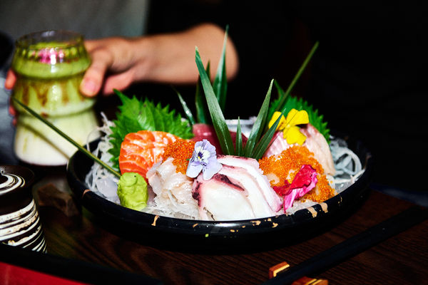 a black bowl holds a selection of sashmin, rice noodles, fish rowe and decorated in a tropical fashion. There is wabisabi on the side of the bowl. Out of focus behind a hand holds a pretty glass of a matcha drink.