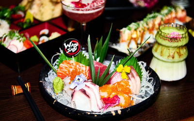 A group shot of plated of sushi and sashimi. Theres is a bento box with sushi items inside. There are two cocktails in shot, one green matcha, the other and elegant red cocktail.