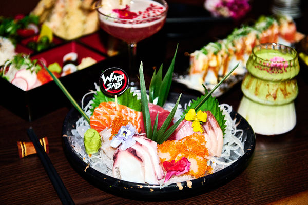 A group shot of plated of sushi and sashimi. Theres is a bento box with sushi items inside. There are two cocktails in shot, one green matcha, the other and elegant red cocktail.