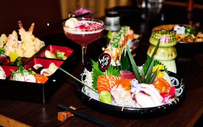 A close-up of an elegantly arranged Japanese-style meal on a dark wooden table. In the foreground is a round black plate filled with assorted sashimi (slices of raw fish and seafood) nestled on a bed of shredded daikon. Garnishes include a green leaf, a small mound of wasabi, tiny edible flowers, and bright orange fish roe. A small sign labeled “Wabi Sabi” is inserted among the slices of fish. Chopsticks rest on a chopstick holder next to the plate. In the midground, there’s a tall, pinkish cocktail in a coupe glass garnished with petals, and beyond that, more trays and bowls of sushi rolls, tempura, and side dishes can be seen. The background is softly out of focus, giving emphasis to the vibrant food presentation.