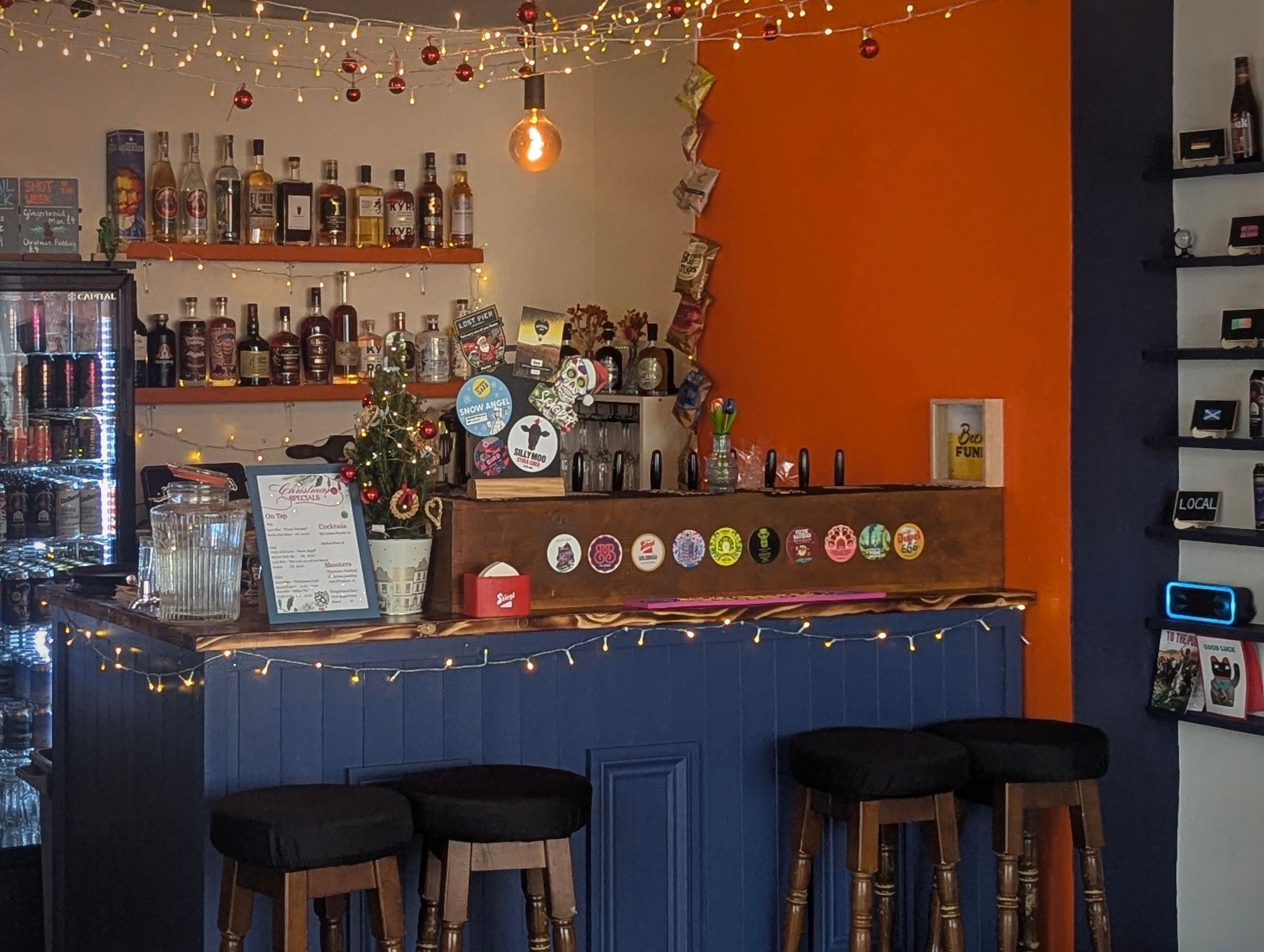 An interior of a bar. It has orange walls and a dark blue bar with barstools. Many beers line shelves on the walls and there are bar stools. There are fairly lights festooned over the ceiling.