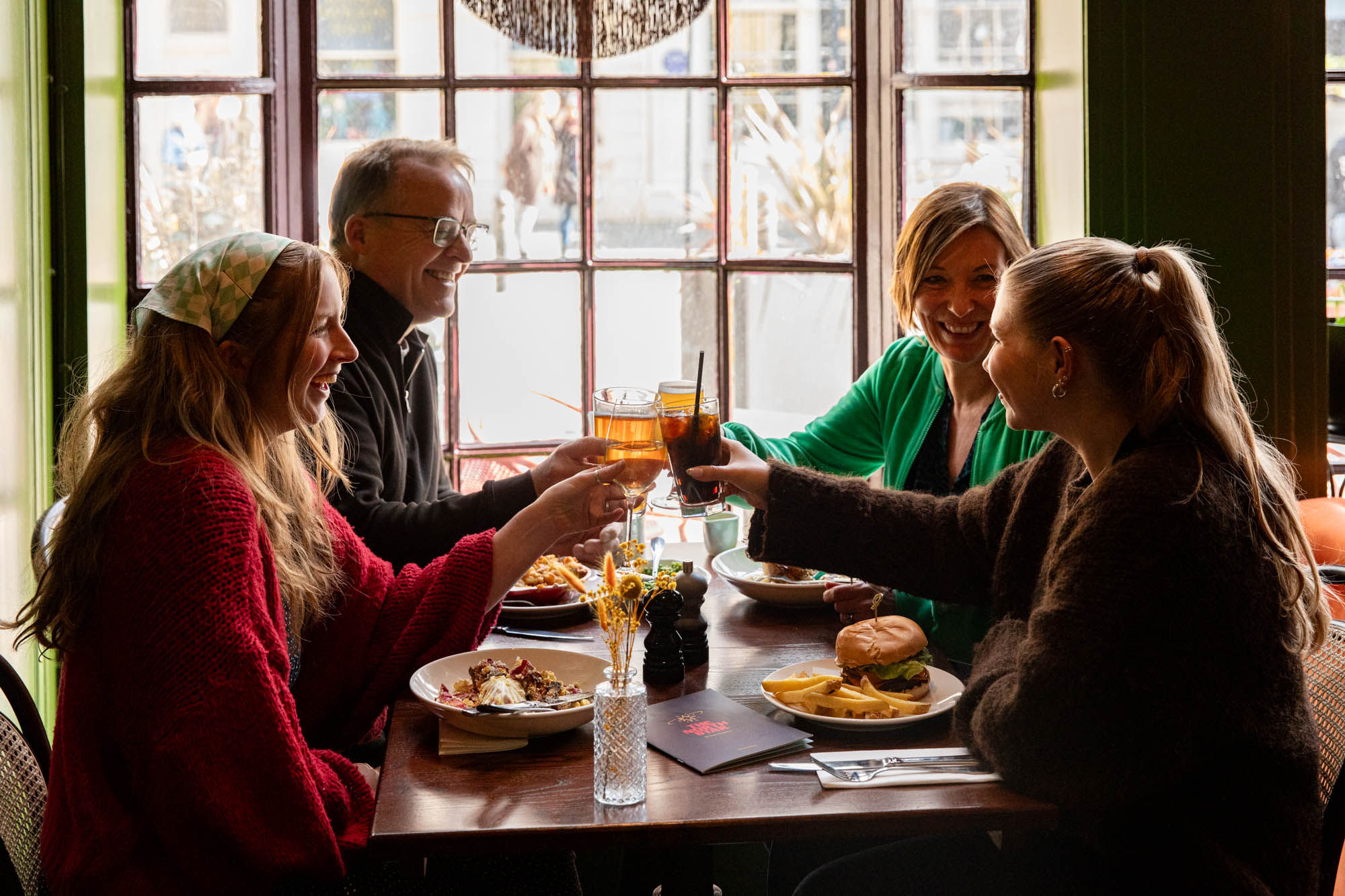 group of friends sitting at the table next to window and enjoying food and drink