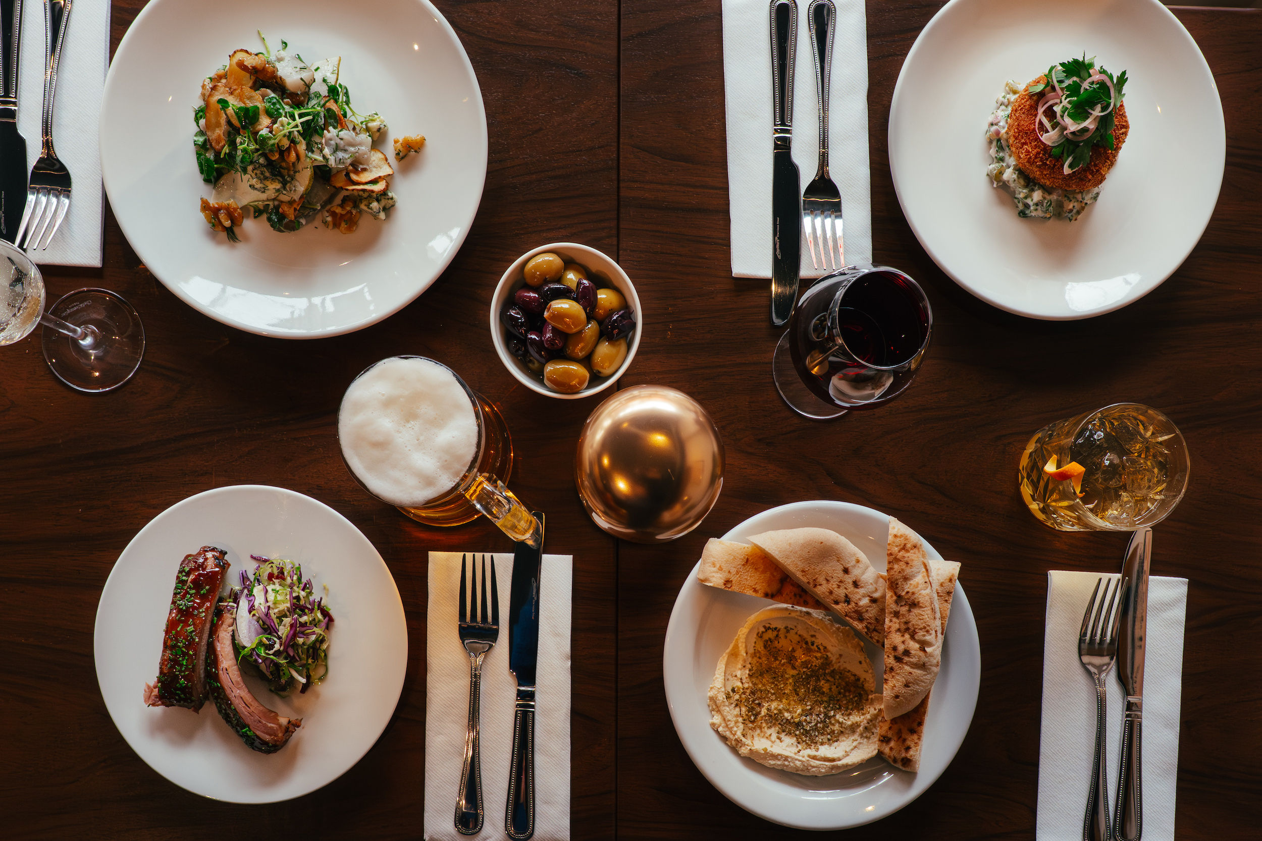 a shot from above of the dishes at The North Star Brighton. Including ribs, hummus, fishcakes, olives and beer. a picture from above looking down onto a wooden table. 