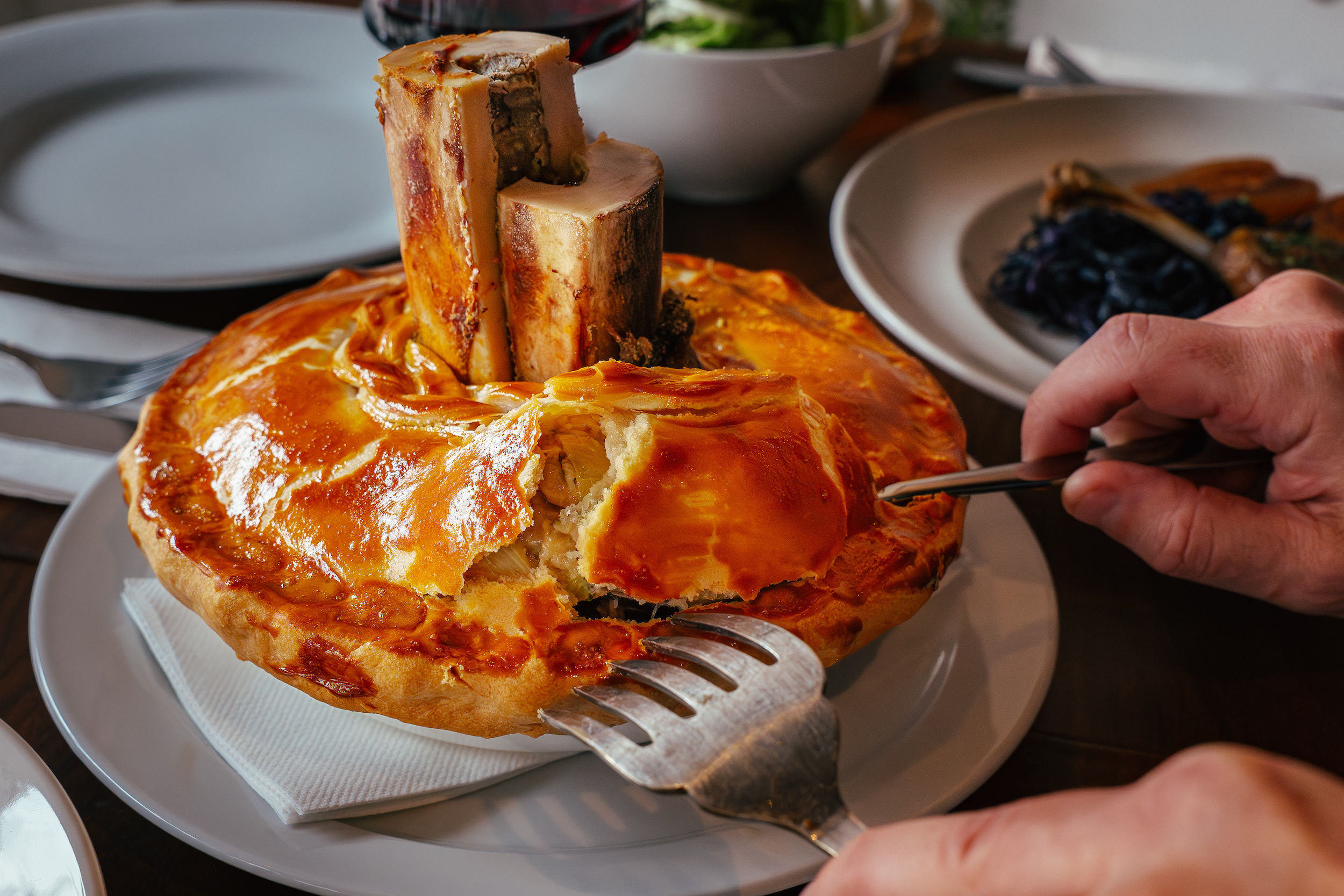 Steak pie with a gorgeous golden pastry and the bone marrow poking out. With a diner approaching the dish with knives and forks. 