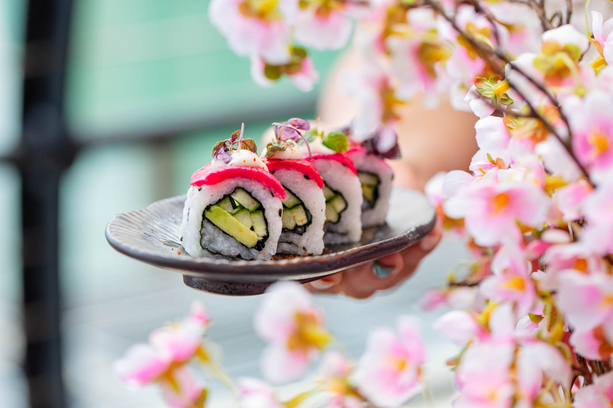 a set of 4 sushi rolls on a decorative plate is being held next to a pink cherry blossom tree.