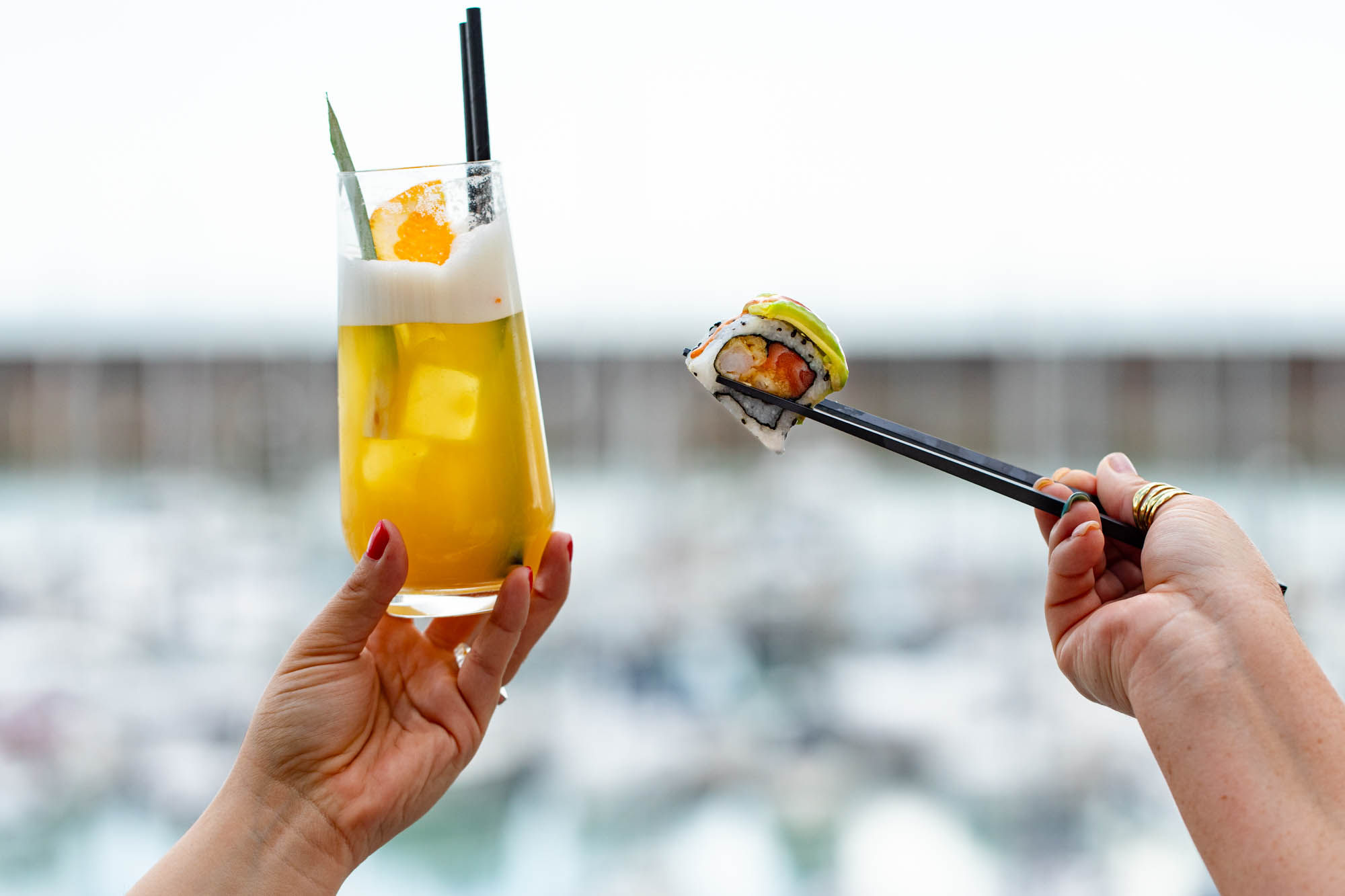 in focus we saee one hand holding up an orange cocktail drink in a tall glass. The other has a piece of sushi held by chopsticks. The background is blurred out