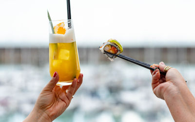 a cocktail is being held in one hand, and a sushi roll in the other. The background is the marina but it is blurred out.