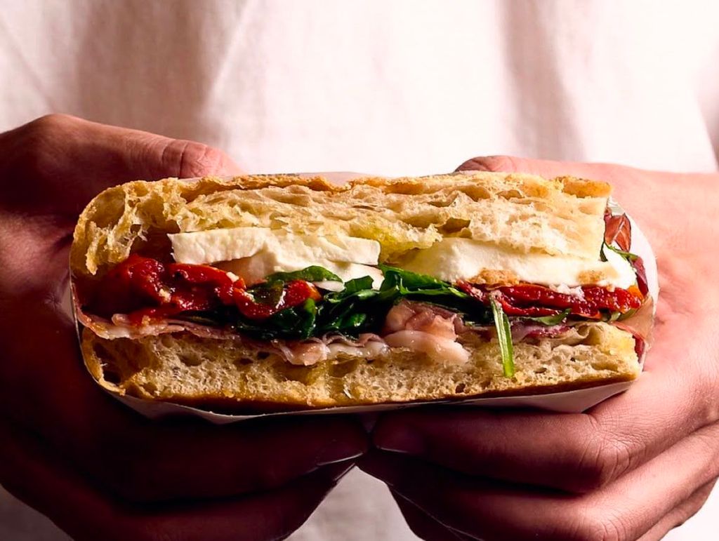 A large foccacia sandwich with fresh ingredients being held in front of the camera against a white t-shirt.