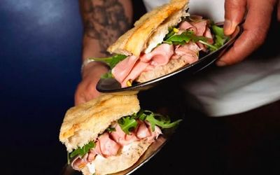 Ham sandwiches with fresh rocket with foccacia bread on black plates being served by a man in a white t-shirt