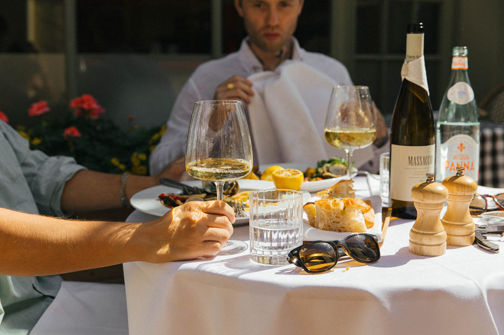 Two men alfresco dining on a sunny day with glasses of white wine and a table of Italian food.