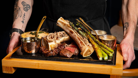 A meat platter on a slate board being held by a chef in a black apron with tattoos on arm.