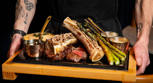 A meat platter on a slate board being held by a chef in a black apron with tattoos on arm.
