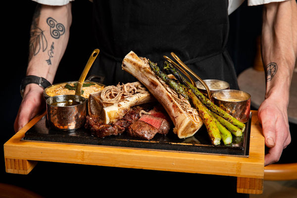 A meat platter on a slate board being held by a chef in a black apron with tattoos on arm.