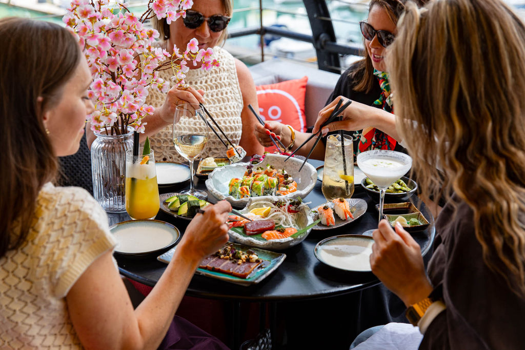 A group of 4 dining on sushi in an outdoor setting. Sat round a table with many sushi dishes and drinks on it. All are holding chopsticks.