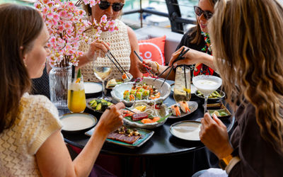 A group of 4 dining on sushi in an outdoor setting. Sat round a table with many sushi dishes and drinks on it. All are holding chopsticks.