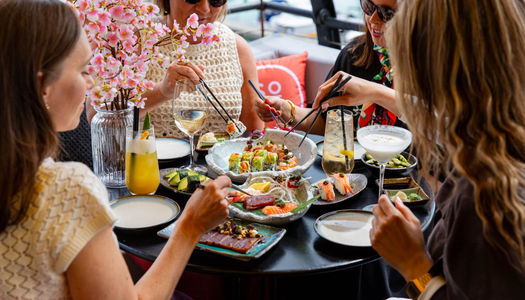 A group of 4 dining on sushi in an outdoor setting. Sat round a table with many sushi dishes and drinks on it. All are holding chopsticks.