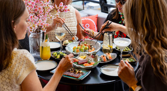 A group of 4 dining on sushi in an outdoor setting. Sat round a table with many sushi dishes and drinks on it. All are holding chopsticks.