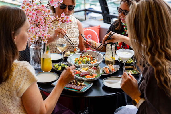 A group of 4 dining on sushi in an outdoor setting. Sat round a table with many sushi dishes and drinks on it. All are holding chopsticks.