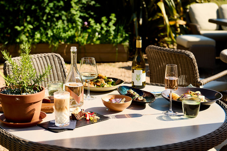 An alfresco summer feast with wine on the terrace with foliage in the background.