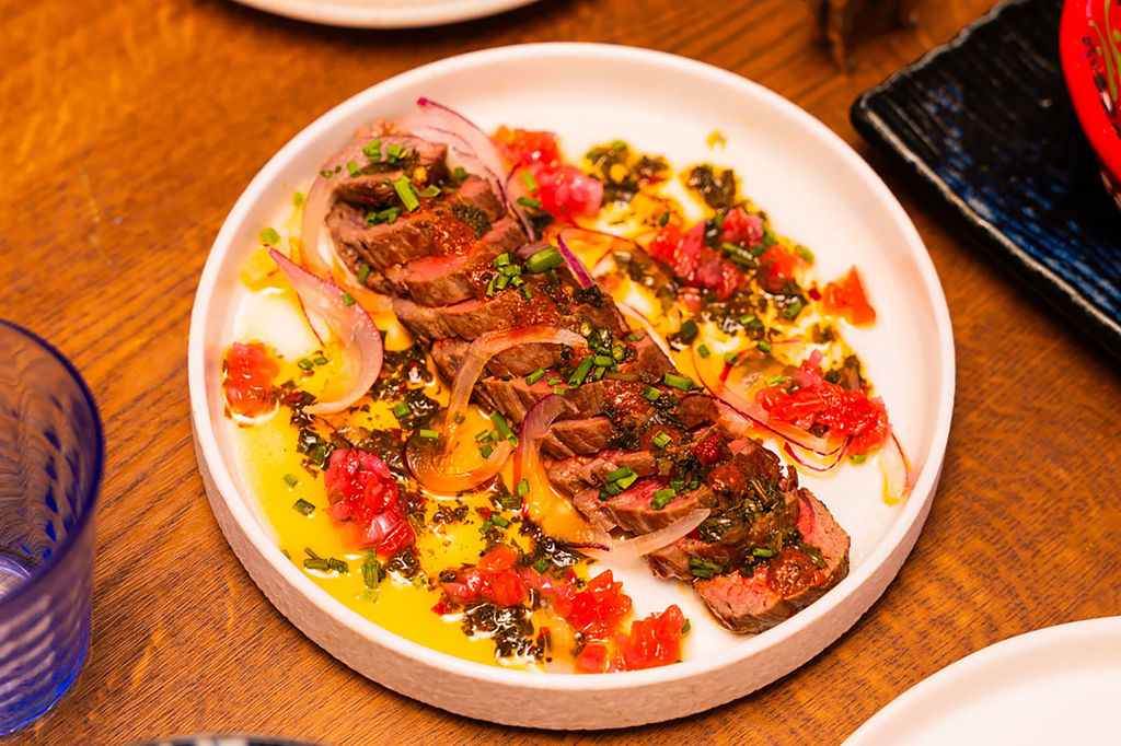 A close up photo of a large white shallow dish with thinly sliced meat, sliced red onions and tomatoes.