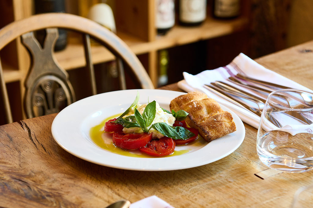 A white plate with mozzarella, sliced tomatoes and focaccia with fresh basil on a wooden table next to a chair.