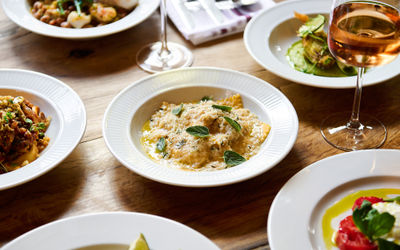 A wooden table with lots of white plates full of Italan dishes such including pasta