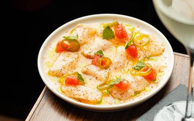 A close-up shot of a round white plate presenting thin slices of raw white fish arranged in a light citrus sauce. The dish is garnished with bright red grapefruit segments, sliced pickled jalapeños, fresh mint leaves, and a sprinkle of reddish chili seasoning. A stemware glass rests partially in the foreground.