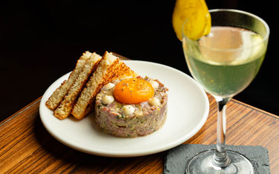 A white plate featuring a neatly molded round stack of seasoned steak tartare topped with small dollops of cream and a vibrant raw egg yolk lightly dusted with black pepper.