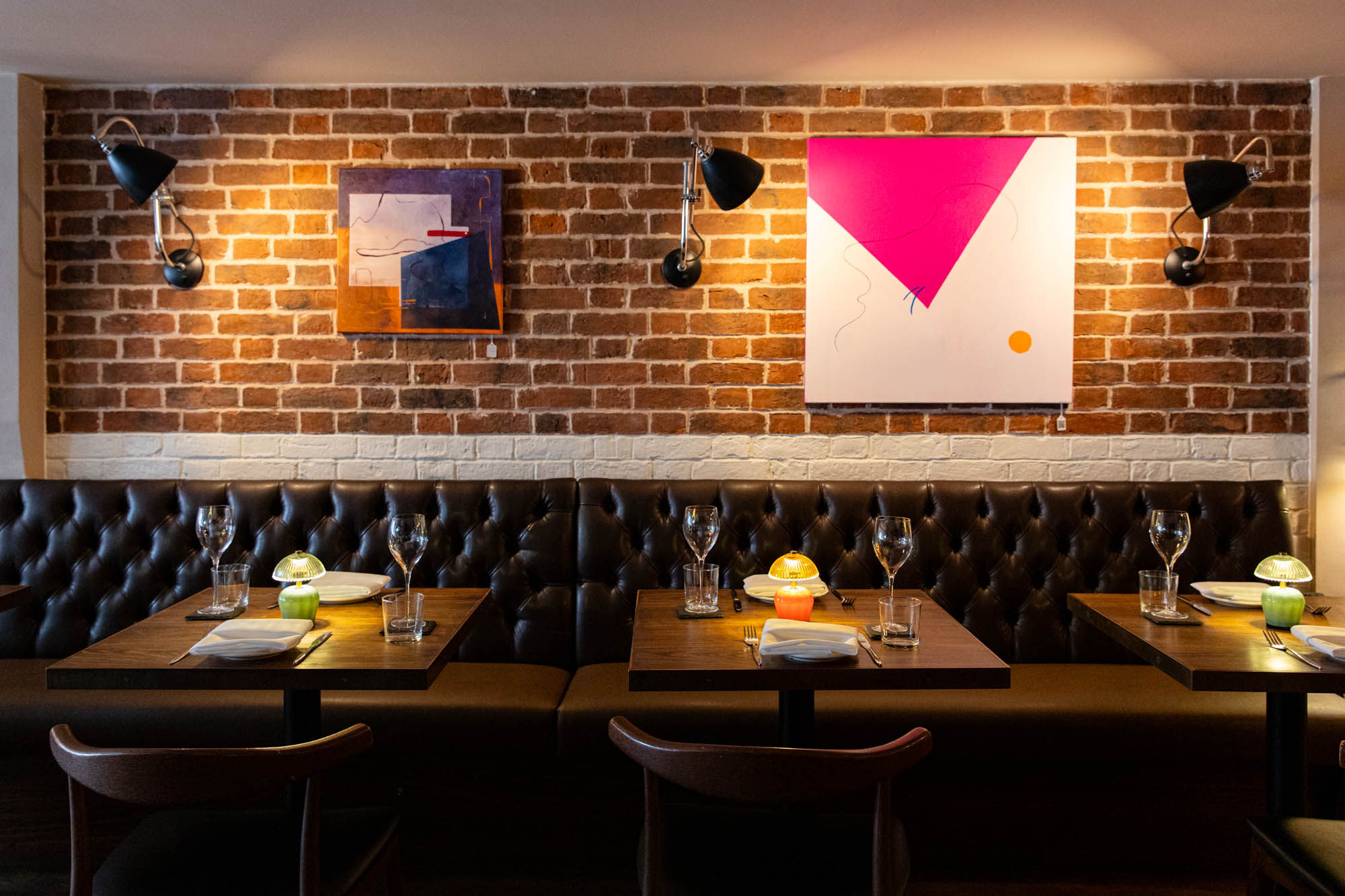 An warm, cozy restaurant dining space featuring a dark brown tufted leather banquette running along an exposed red brick wall