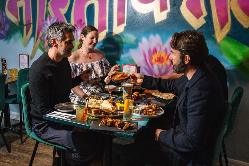 A group of friends eating around a table full of Indian food with drinks 