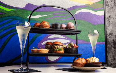 a picture of an afternoon tea on a tow tiered plat. The background is colourful and there are two glasses of sparkling wine on the table.