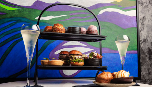 a picture of an afternoon tea on a tow tiered plat. The background is colourful and there are two glasses of sparkling wine on the table.
