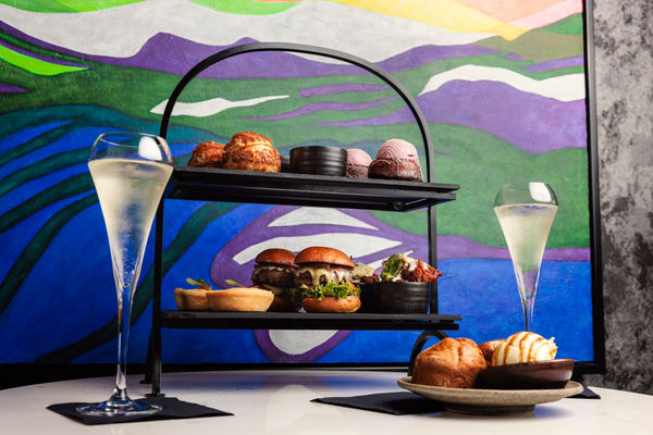 a picture of an afternoon tea on a tow tiered plat. The background is colourful and there are two glasses of sparkling wine on the table.