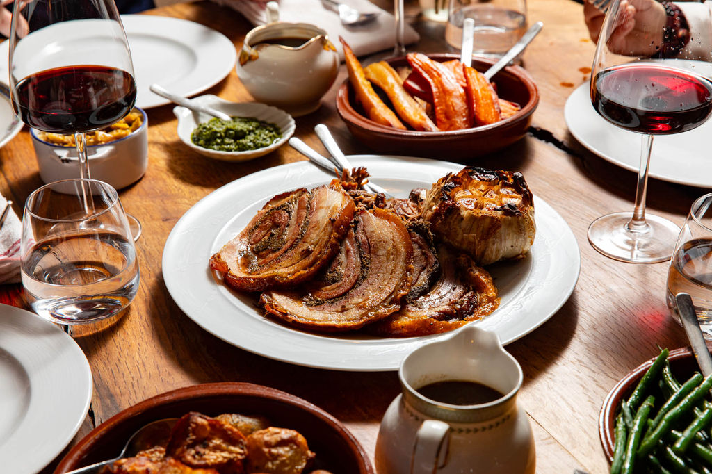Sunday roast laid out on a table with glasses of red wine and a plate in the middle of the table with sliced meat.