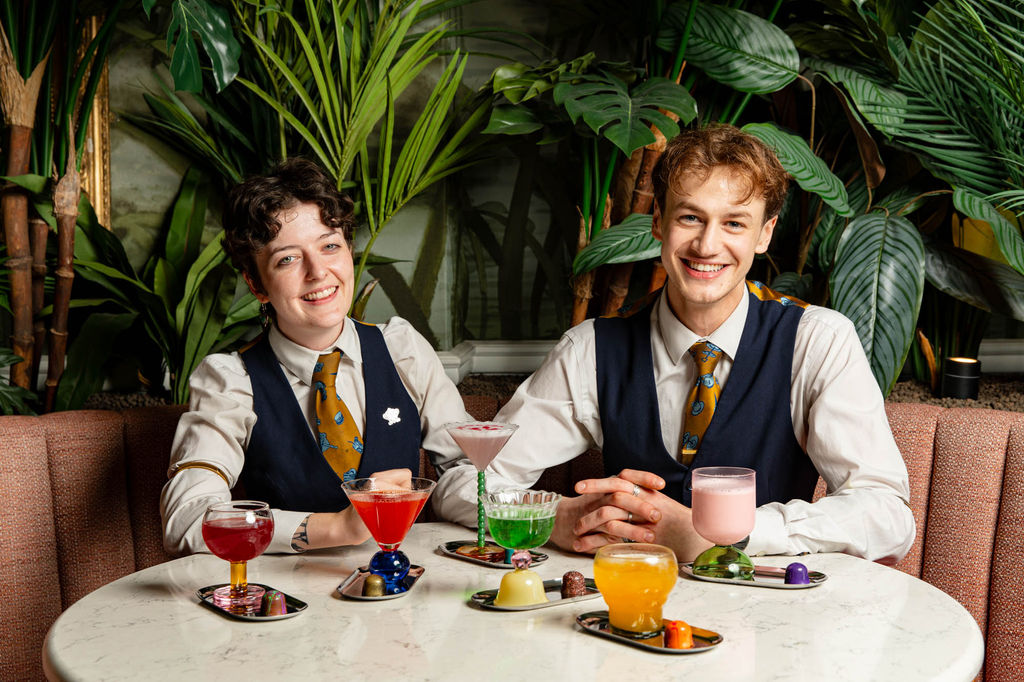 Two people sat at at table with a collection of colourful cocktails and plants behind them