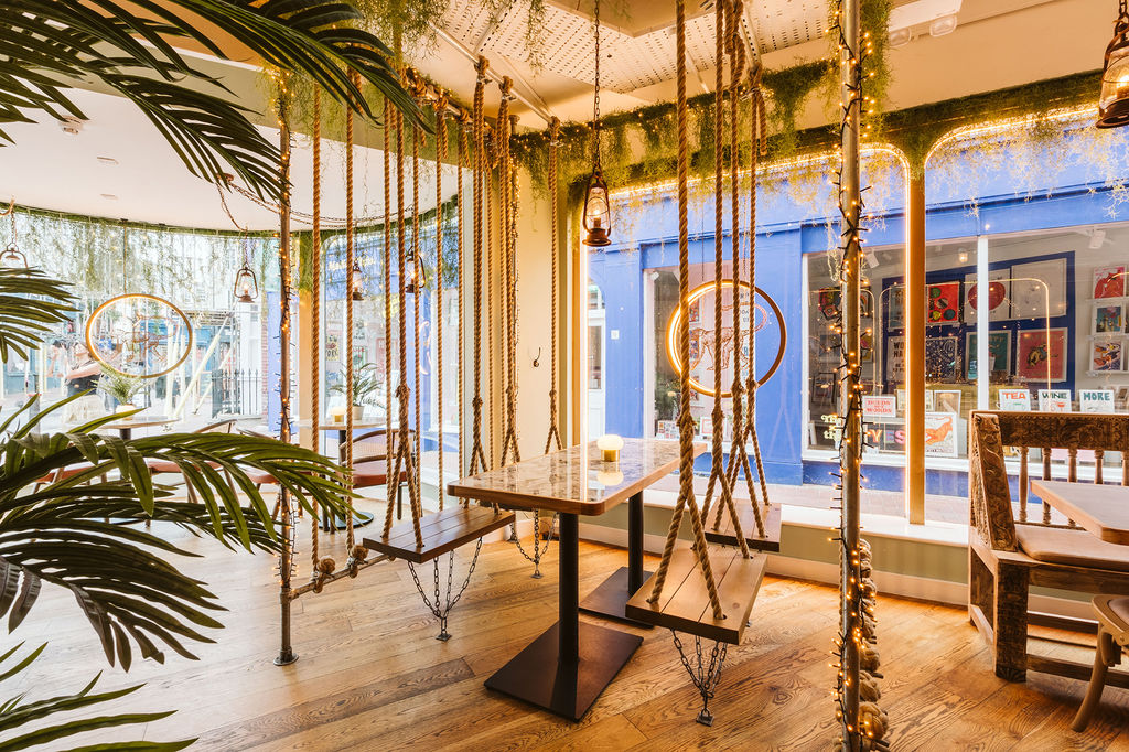 A swing seat and table in the window of Indian restaurant with plants and ambient lighting.