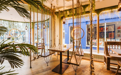 A swing seat and table in the window of Indian restaurant with plants and ambient lighting.