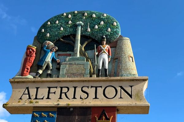The carved wood village sign of Alfriston depicting a scene in their market square is pictured against a bright blue sky
