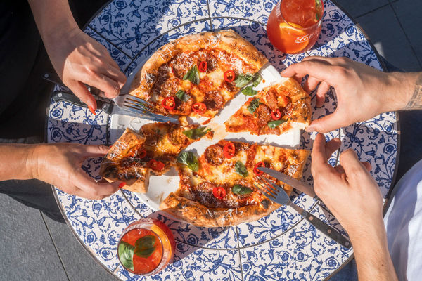Overhead photo of a sliced pizza on a table with a blue and white pattern, being eaten by two people with a Negroni drink