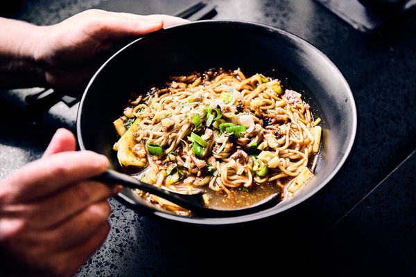 A large black bowl of noodles in a broth with hands holding the bowl and a spoon