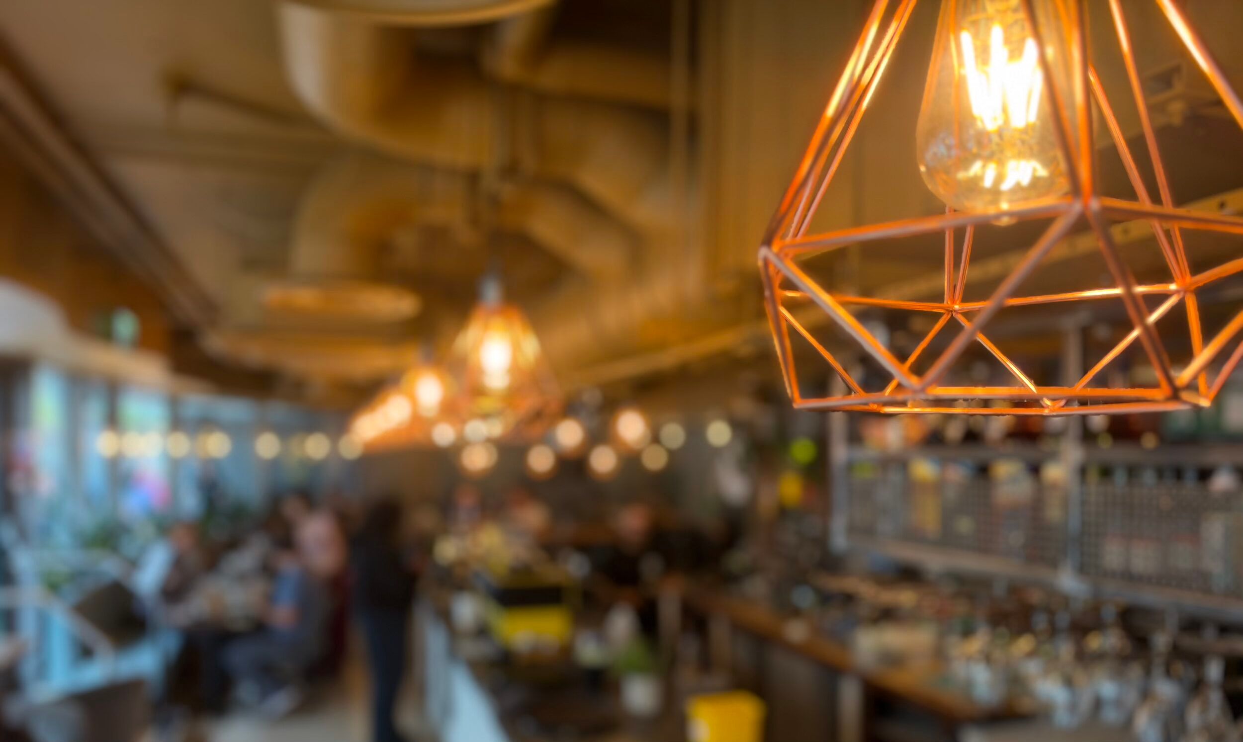 Copper geometric pendant light with a glowing filament bulb inside the cafe, with the dining room softly blurred behind