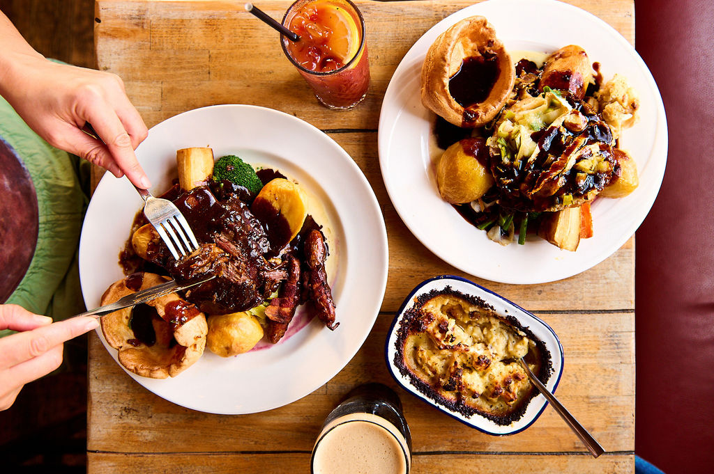 Overhead photo of two plates of Sunday roasts on a wooden table with drinks